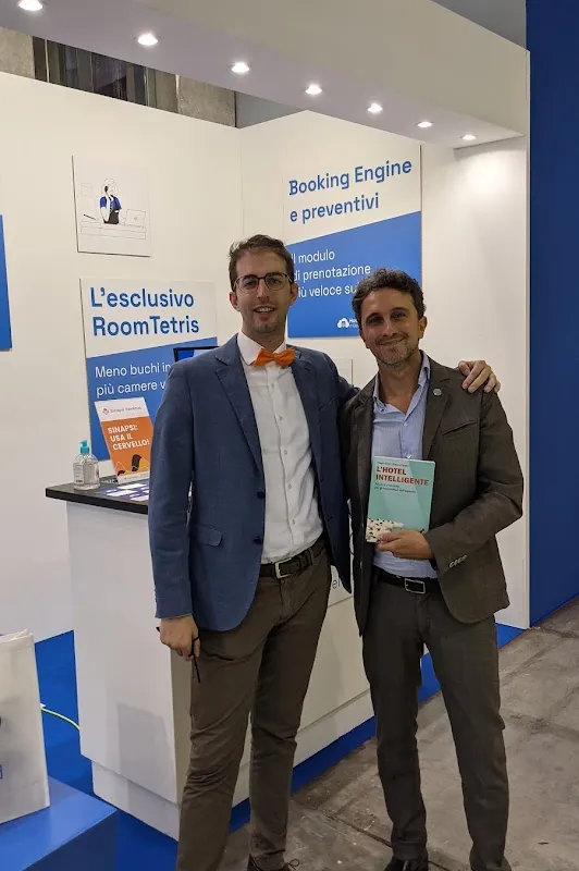 Filippo Battiti allo stand Hotel in Cloud alla Fiera Hotel Bolzano 2021, davanti ai pannelli RoomTetris e Booking Engine, con copia de L'Hotel Intelligente